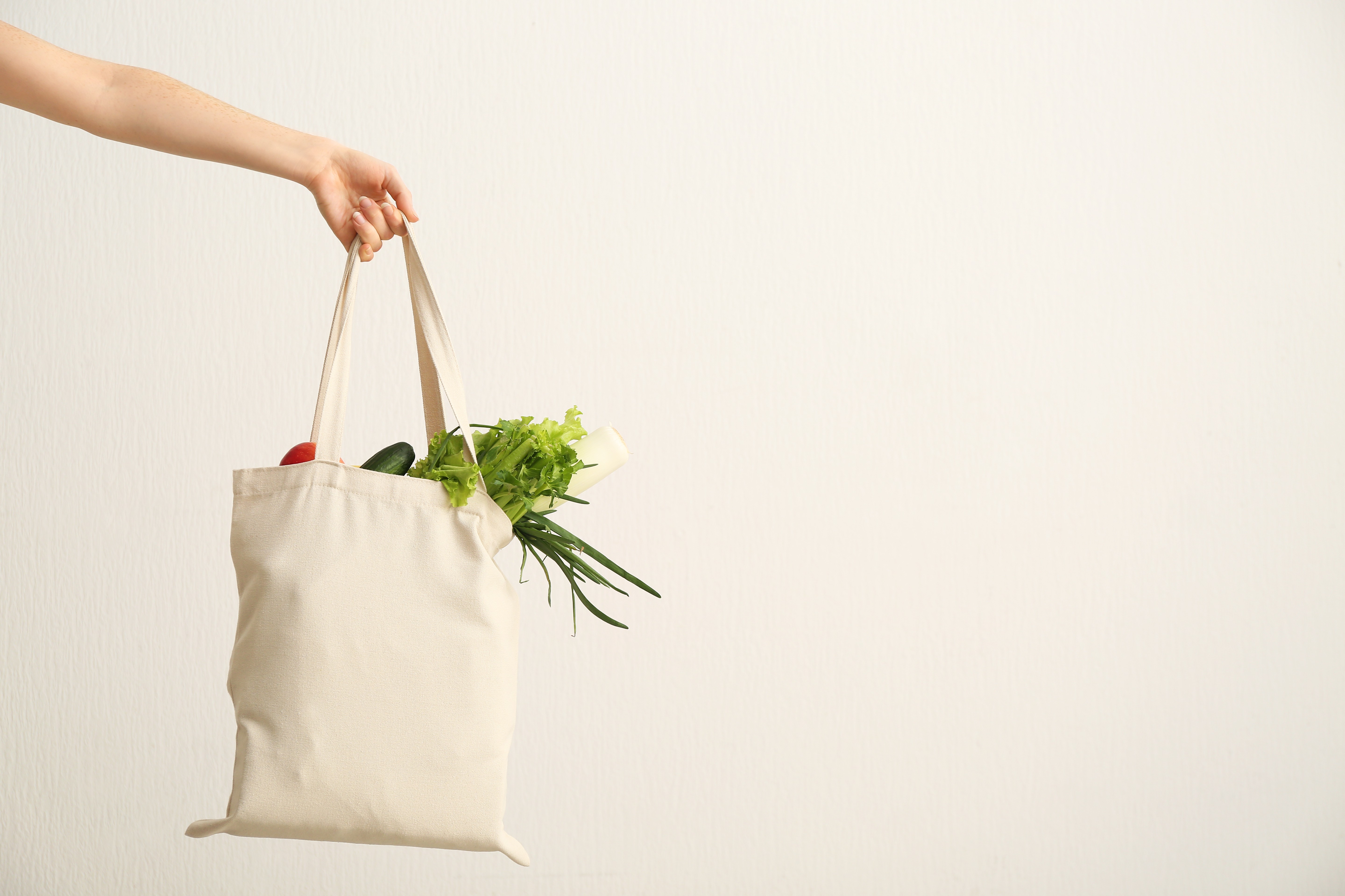 Main tenant un sac en coton rempli de légumes frais contre un mur blanc